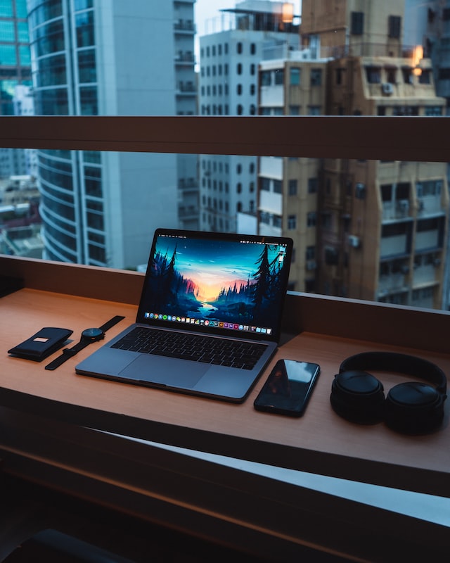 geöffneter und eingeschalteter Laptop auf Schreibtisch. Neben dem Laptop liegen Kopfhörer, ein Handy, eine Hülle und eine Uhr. Laptop befindet sich vor einem Fenster. Im Hintergrund sieht man Hochhäuser.