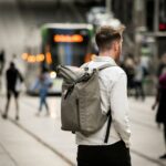 Mann in einem beigen Hemd und einem grauen Rucksack ist von Hinten zu sehen. Im Hintergrund fährt eine Straßenbahn.