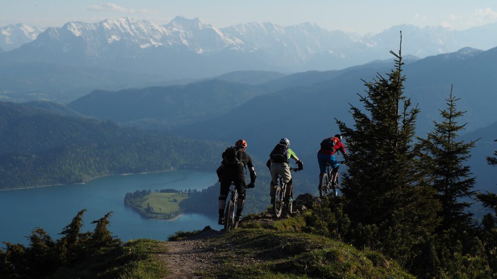 Drei Personen fahren mit dem Mountainbike. Im Hintergrund sind Berge und ein See zu sehen.