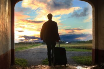 Person steht mit einem Koffer in der Hand und schaut in den Sonnenuntergang.