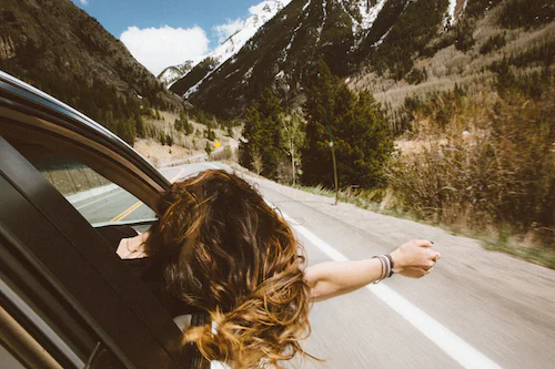 Person lehnt sich aus fahrendem Auto und streckt Arm hinaus. Im Hintergrund sind Berge zu sehen.