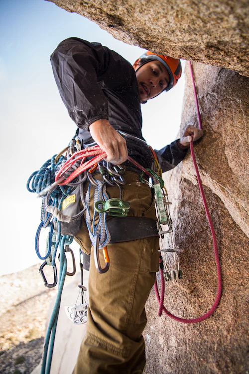 Mann in Kletterausrüstung wie Helm, Karabiner und Seilen steht an einer Felswand und richtet Seile. 