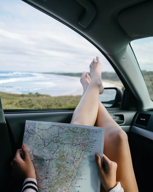Person sitzt im Auto und streckt Füße aus geöffnetem Auto und hält dabei eine Landkarte in der Hand.