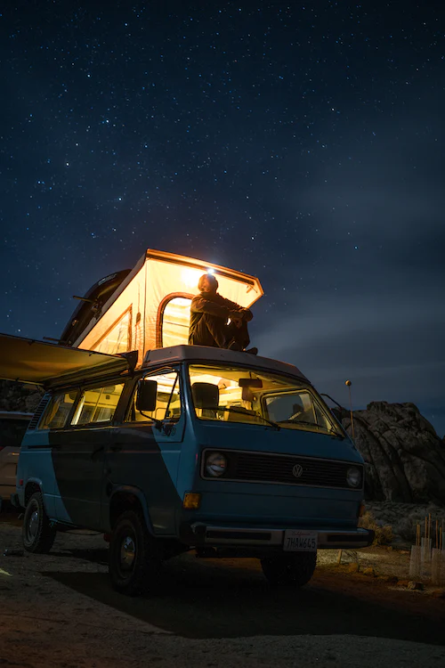 Beleuchteter Van unter Sternenhimmel. Person sitzt auf dem Dach und blickt in den Himmel. 