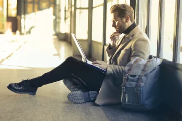 Mann sitzt mit Gepäck am Boden und schaut in einen Laptop