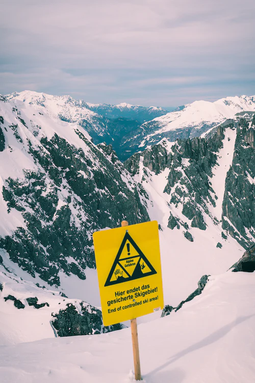 Gelbes Schild mit einem Warnhinweis: "Hier endet das gesicherte Skigebiet" Im Hintergrund ist eine verschneite Berglandschaft zu sehen.