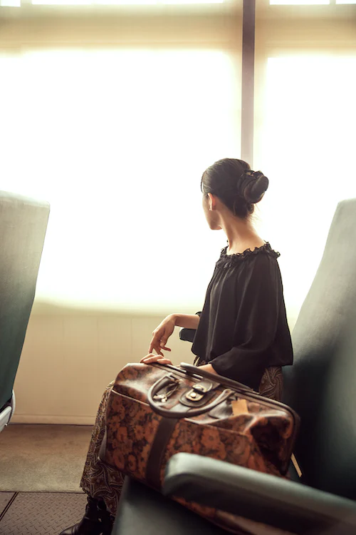 Frau mit Dutt sitzt auf einem Stuhl und blickt aus dem Fenster. Neben ihr steht eine braune Reisetasche.