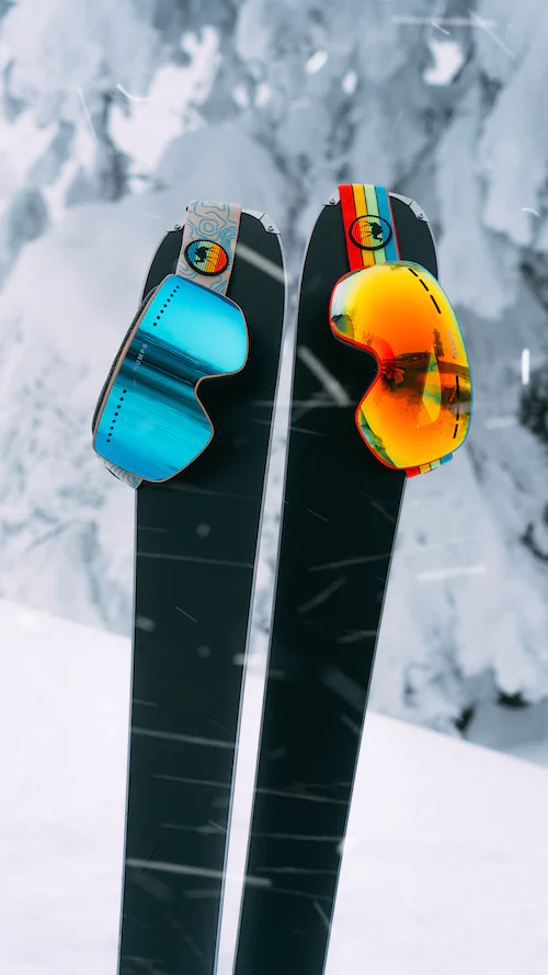 Zwei schwarze in Schnee steckende Skier auf denen eine blaue und eine orange Skibrille hängen.