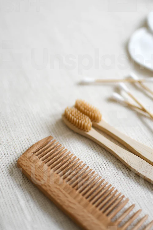 Kamm und  zwei Zahnbürsten aus Holz sowie Wattestäbchen und ein Wattepad liegen auf einem Handtuch. 